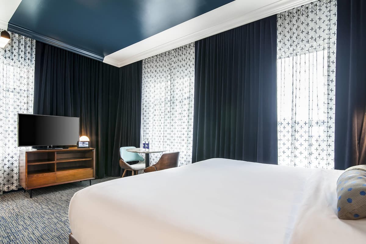 A hotel room with a large bed, a TV on a wooden stand, a small table with a chair, and curtains with a cross pattern.