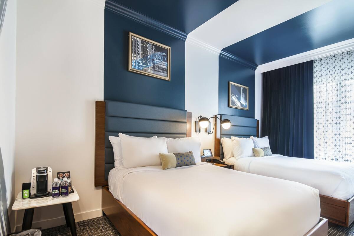 A hotel room with two beds, a nightstand with a phone and a coffee maker, and a small table with a trash bin and a water bottle.
