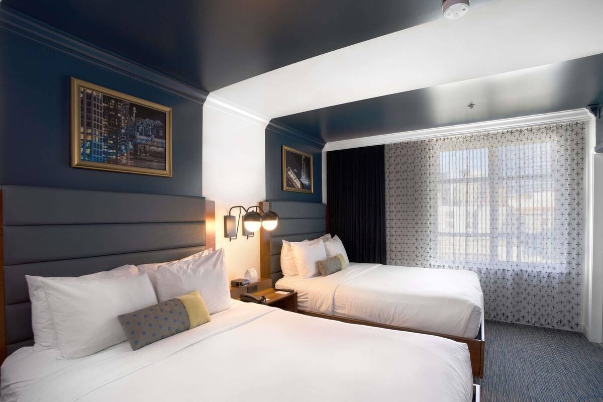 A hotel room with two beds, a nightstand, a wall-mounted lamp, a window with curtains, and framed pictures on the wall.