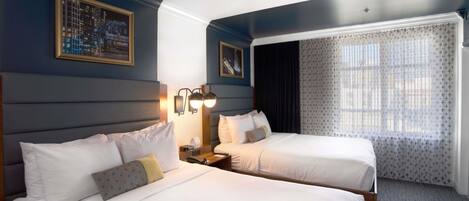 A hotel room with two beds, a nightstand, a wall-mounted lamp, a window with curtains, and framed pictures on the wall.