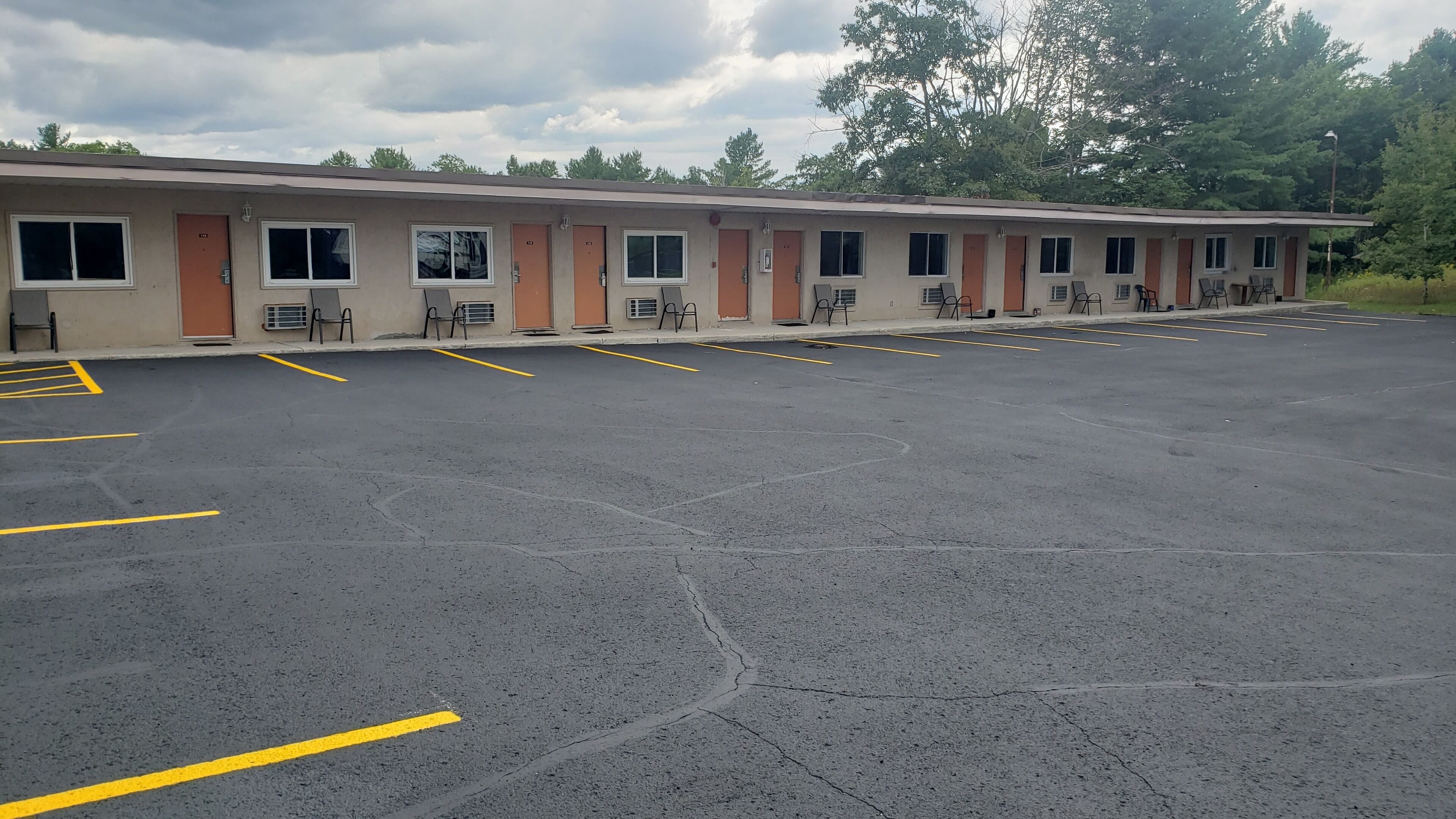 Un motel con varias habitaciones, un estacionamiento y una fila de árboles al fondo.