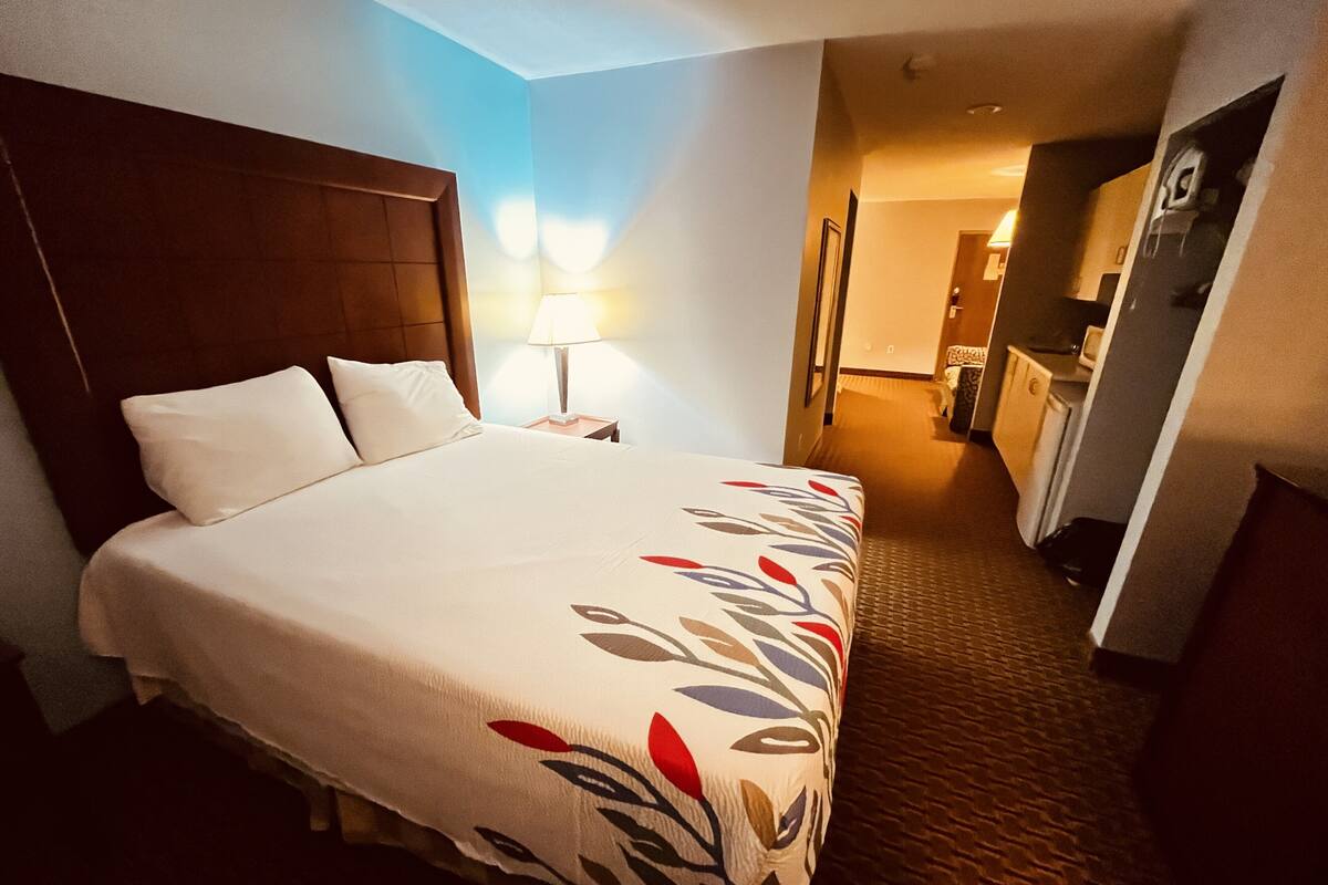 Habitación de hotel con una cama grande, una mesita de noche con lámpara, cabecera de madera y vista al pasillo y al baño.
