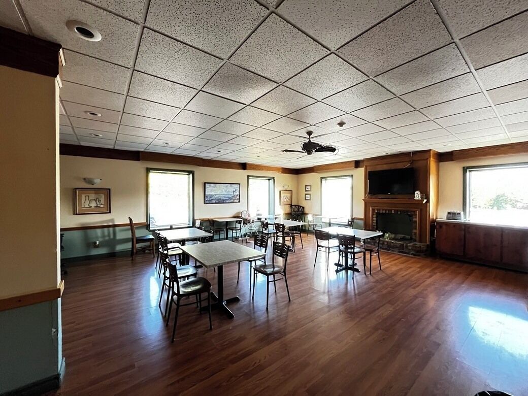A spacious room with wooden flooring, multiple tables and chairs, a fireplace, and a ceiling fan.