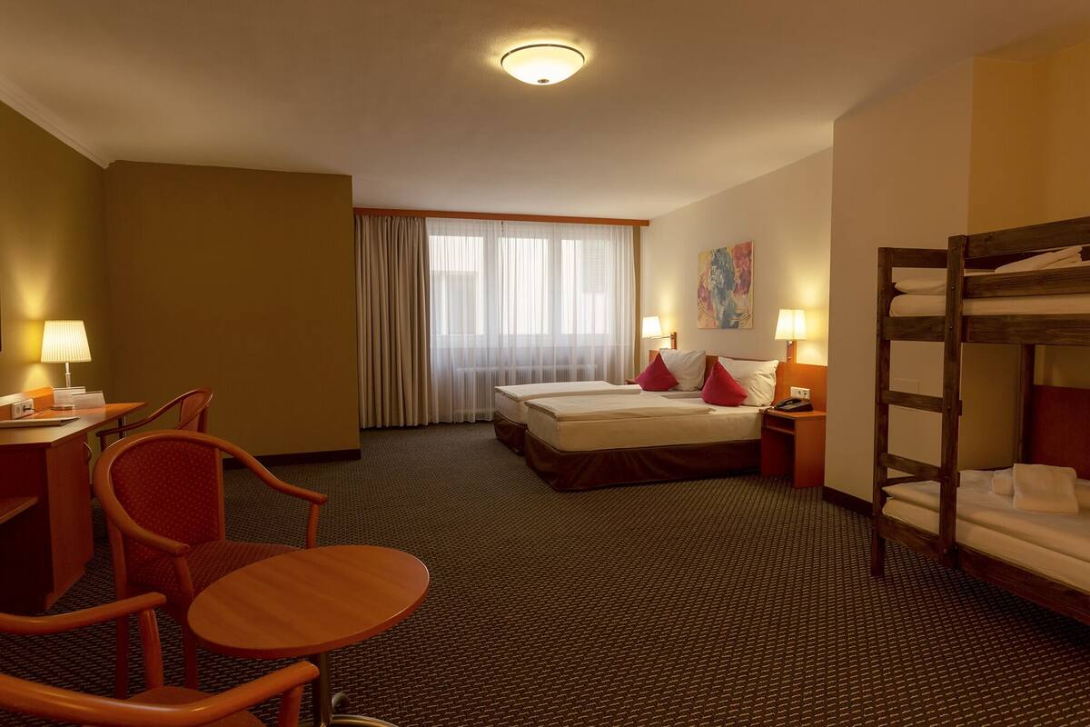 A hotel room with a bed, a desk, a chair, a bunk bed, and a window with curtains.