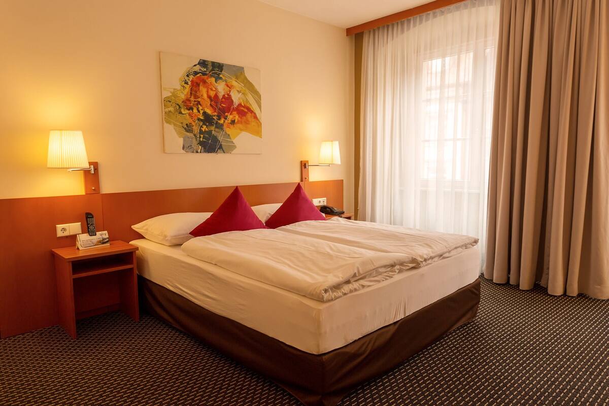 A hotel room with a large bed, two bedside tables, a painting on the wall, and a window with curtains.
