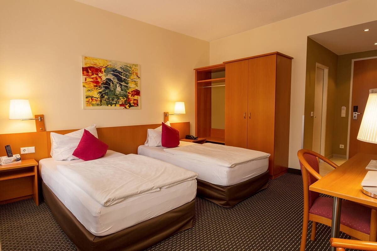 A hotel room with two beds, a desk, a chair, a wardrobe, and a painting on the wall.