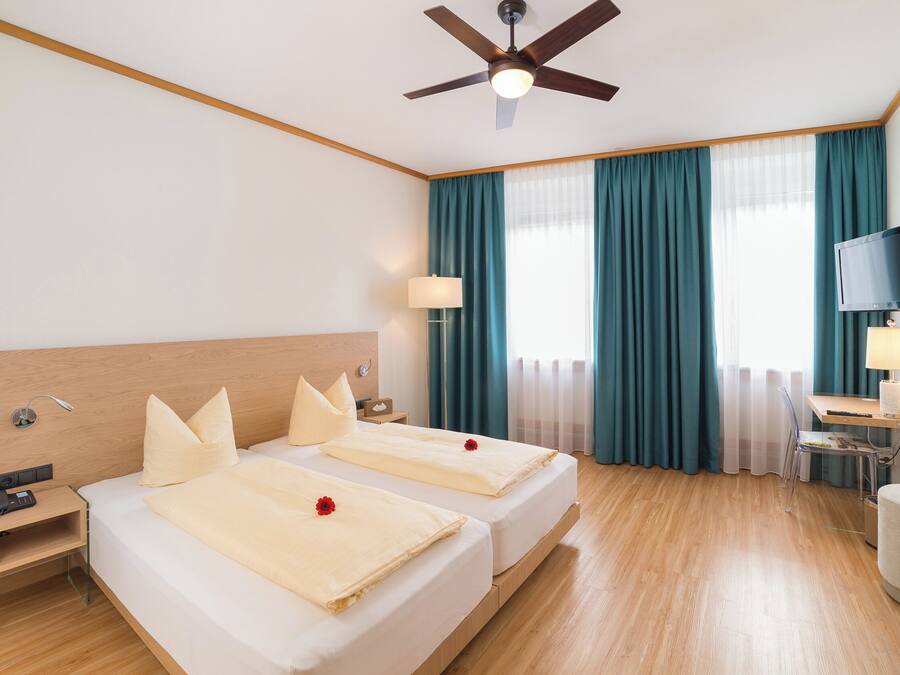 Ein Hotelzimmer mit zwei Betten, einem Schreibtisch mit Stuhl, einem Fernseher und einem Deckenventilator.