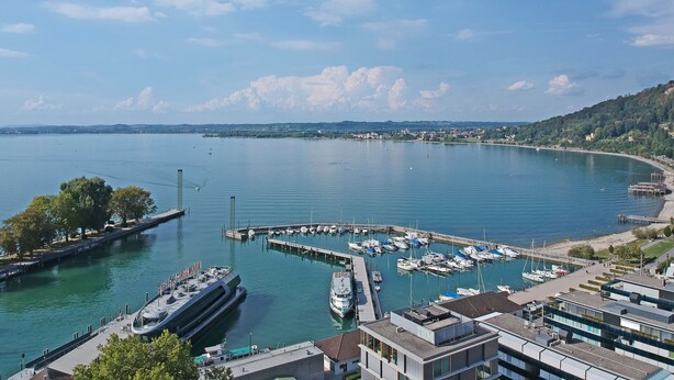 Ein Yachthafen mit vertäuten Booten, ein modernes Gebäude mit Glasfassade und freier Sicht auf Wasser und entfernte Hügel.