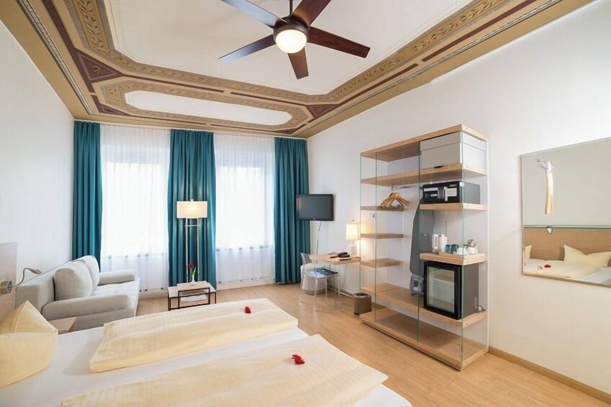 A hotel room with a bed, a TV, a ceiling fan, a glass cabinet, and a mirror.