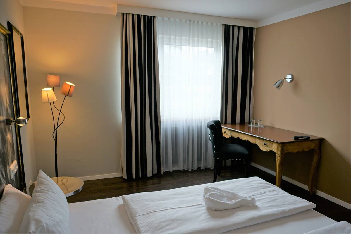 A hotel room with a bed, a desk, a chair, a lamp, and a window with curtains.
