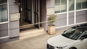 La entrada del Hotel Calmo en Viena, con una fachada moderna, un toldo rosa y un coche blanco estacionado al frente.