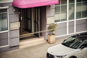 De ingang van Hotel Calmo in Wenen, met een modern gevelontwerp, een roze luifel en een witte auto die ervoor geparkeerd staat.