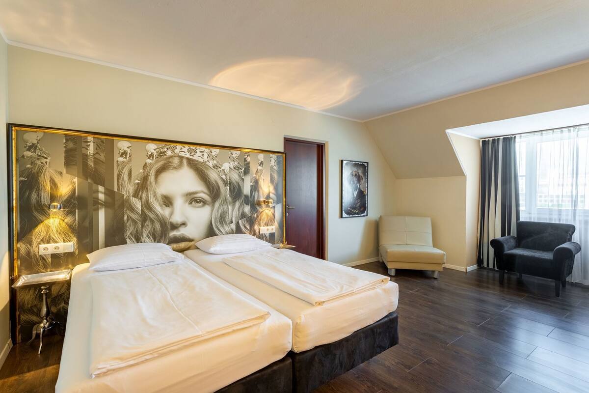 A hotel room with a large bed, a chair, a desk, and a large poster on the wall.