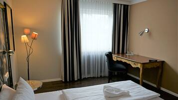 A hotel room with a bed, a desk, a chair, a lamp, and a window with curtains.