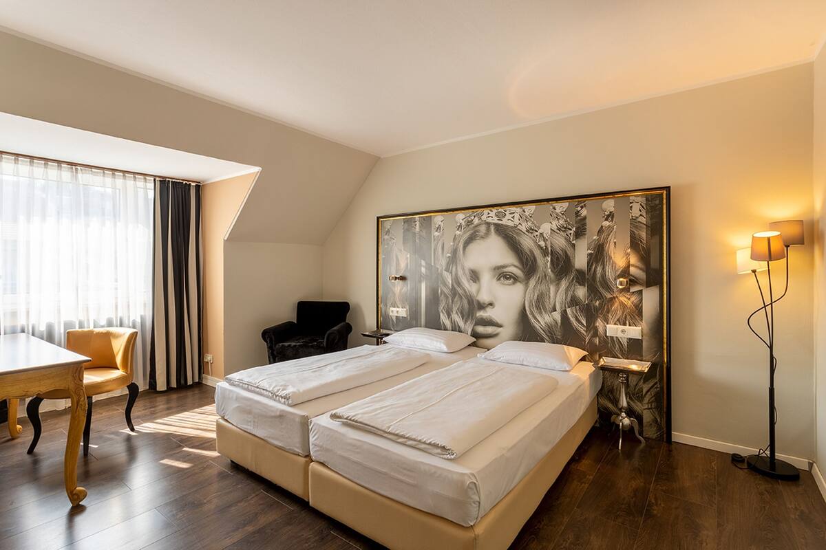 A hotel room with a large bed, a desk, and a chair. There is a painting on the wall and a lamp on the side table.