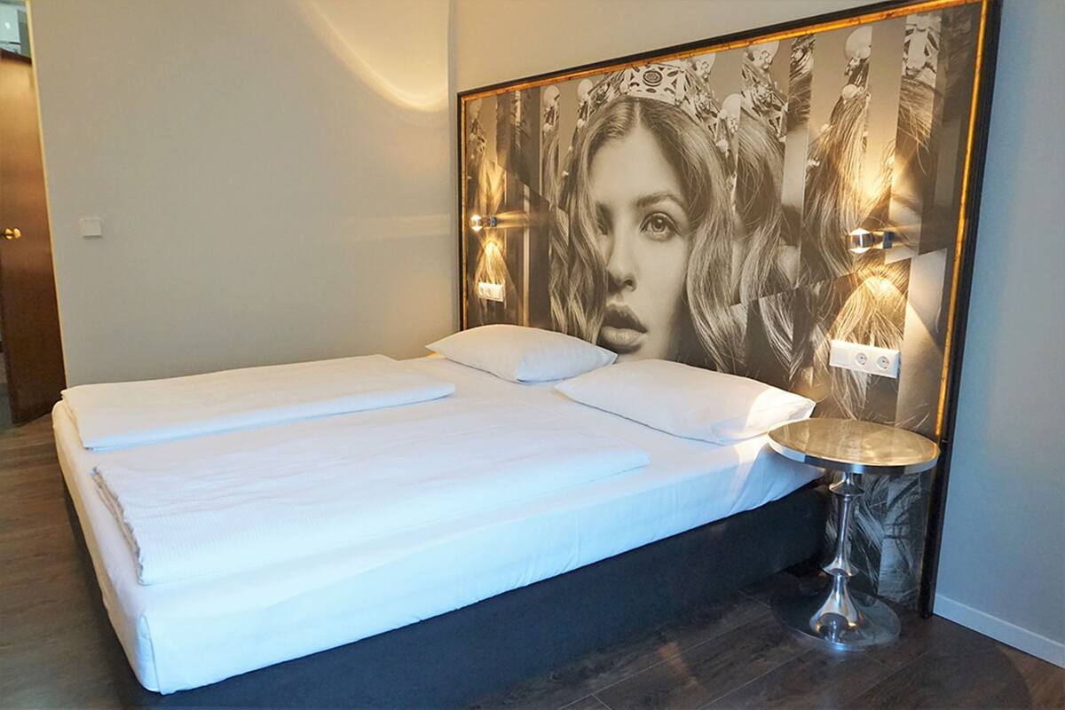 A neatly made bed with white linens and pillows, a headboard with a large framed picture, and a small round table with a lamp beside the bed.