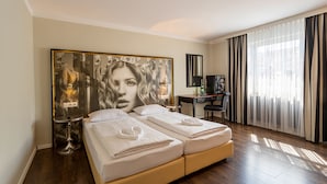 Habitación de hotel con una cama grande, un escritorio y una silla. Hay un cuadro en la pared, una ventana con cortinas y un televisor.