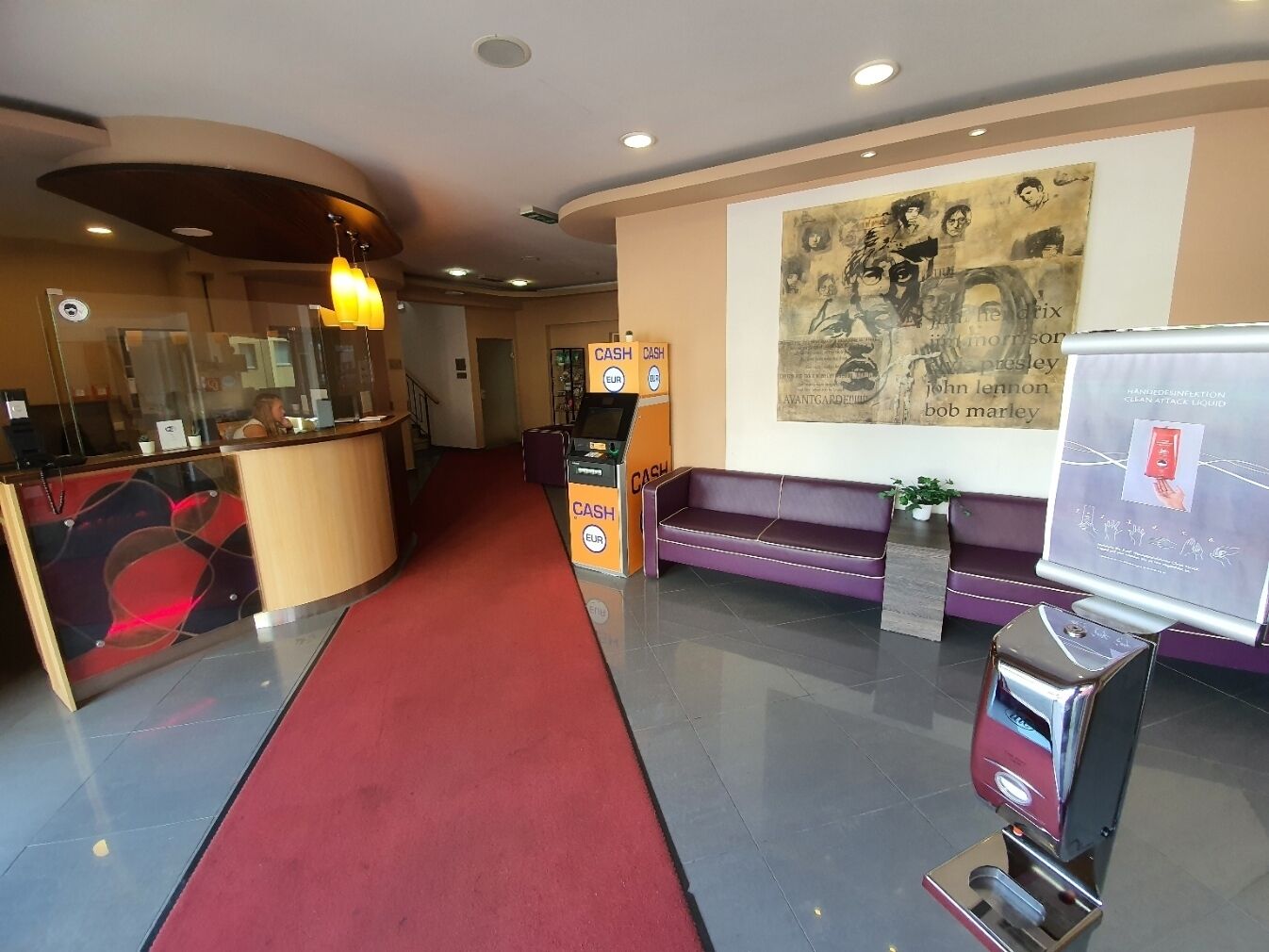 Um saguão de hotel com uma mesa de recepção, um tapete vermelho, um sofá, um cartaz e um dispensador de álcool em gel.