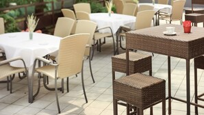 An outdoor patio area with round tables and wicker chairs, surrounded by greenery. - Arion Hotel Vienna Airport (Schwechat)