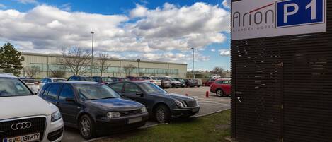 Un área de estacionamiento con varios coches, un cartel que indica 'Airport Hotel P1' y un edificio al fondo.