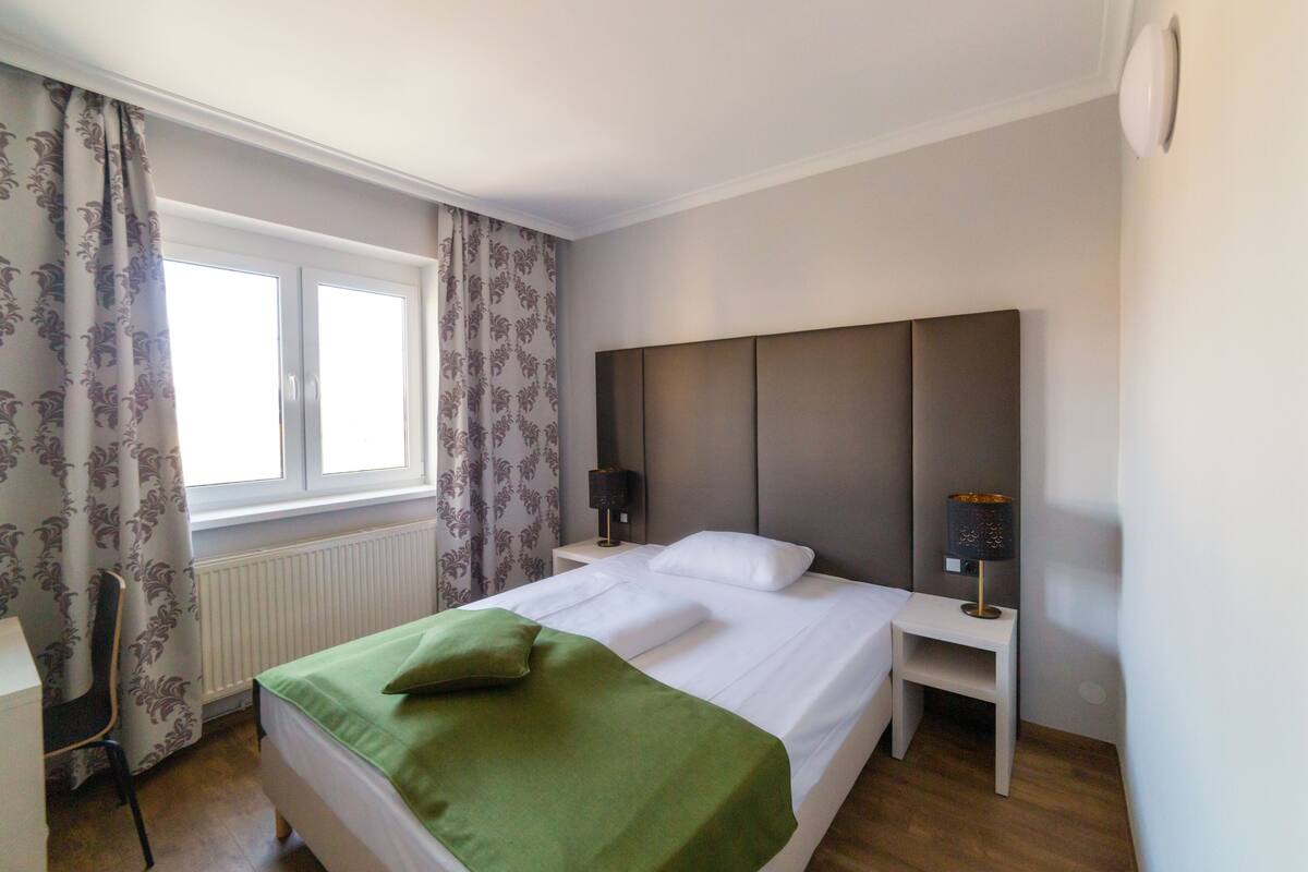 A neatly made bed with a green blanket and pillows, a bedside table with a lamp, a chair, and a window with curtains.