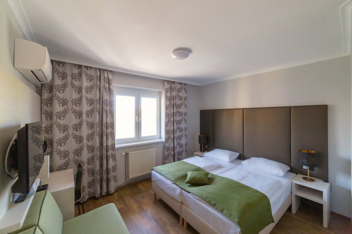 A hotel room with a bed, a TV, a desk, and a window with curtains.