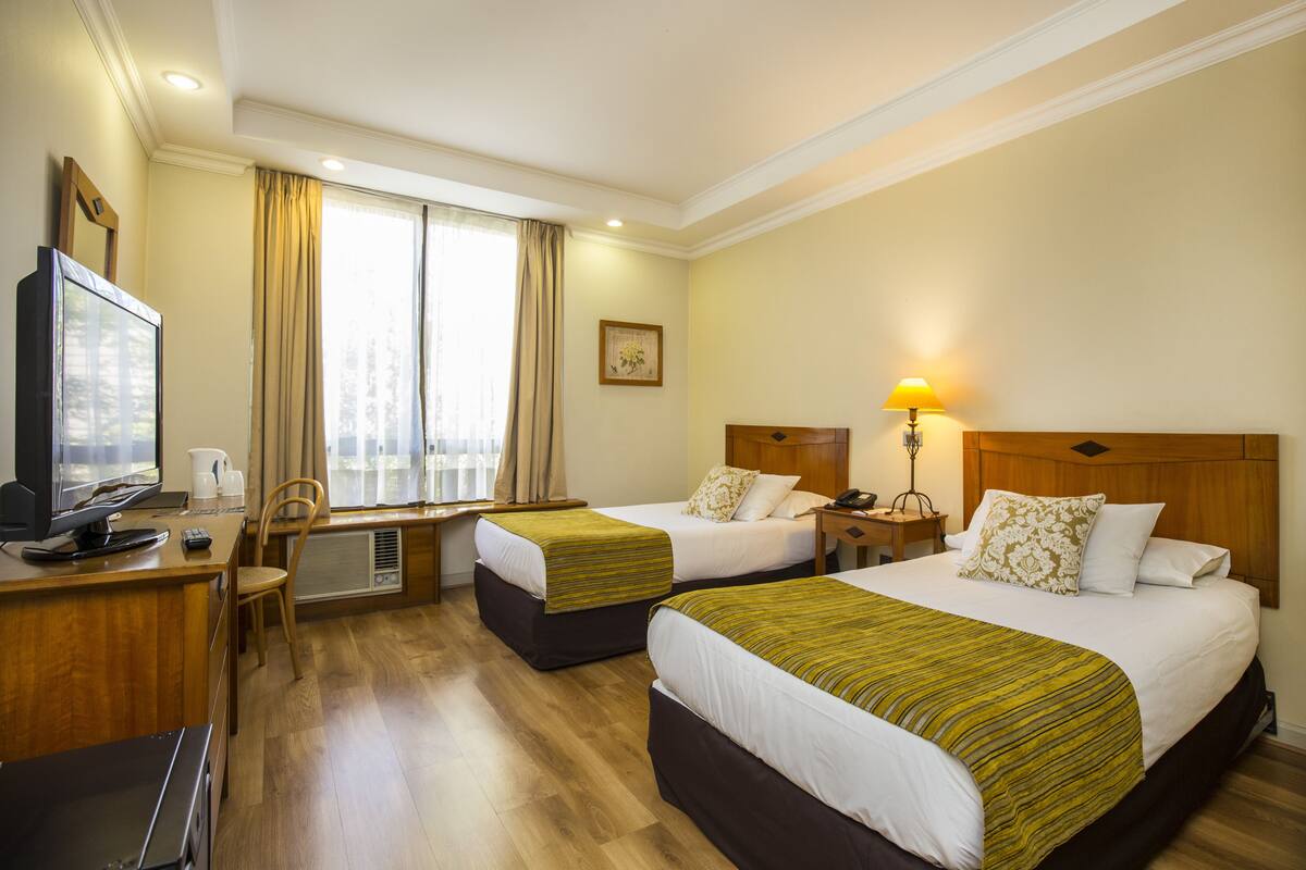 Une chambre d’hôtel avec deux lits, un bureau avec un ordinateur, une chaise, une fenêtre avec des rideaux et une lampe de chevet.