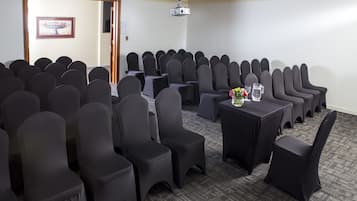 Un salón de conferencias con filas de sillas, un proyector y una mesa con un jarrón de flores.
