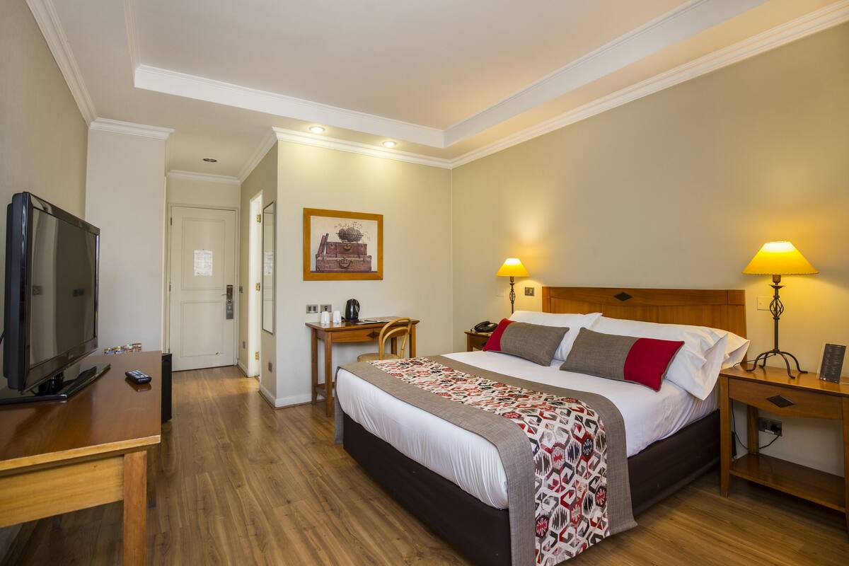 Une chambre d’hôtel avec un lit, des lampes de chevet, un bureau, une télévision et un tableau encadré au mur.