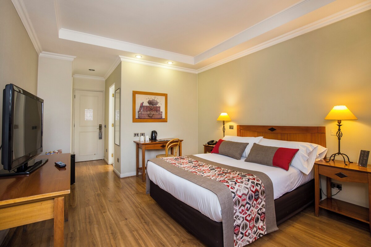 Een hotelkamer met een bed, nachtkastjes, een bureau, een televisie en een deur naar een andere kamer.