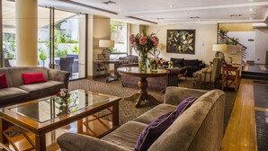 A spacious living area with a sofa, armchairs, a coffee table, and a dining table. There is a large window providing natural light and a view of greenery outside. - Hotel Bonaparte Boutique (Santiago)