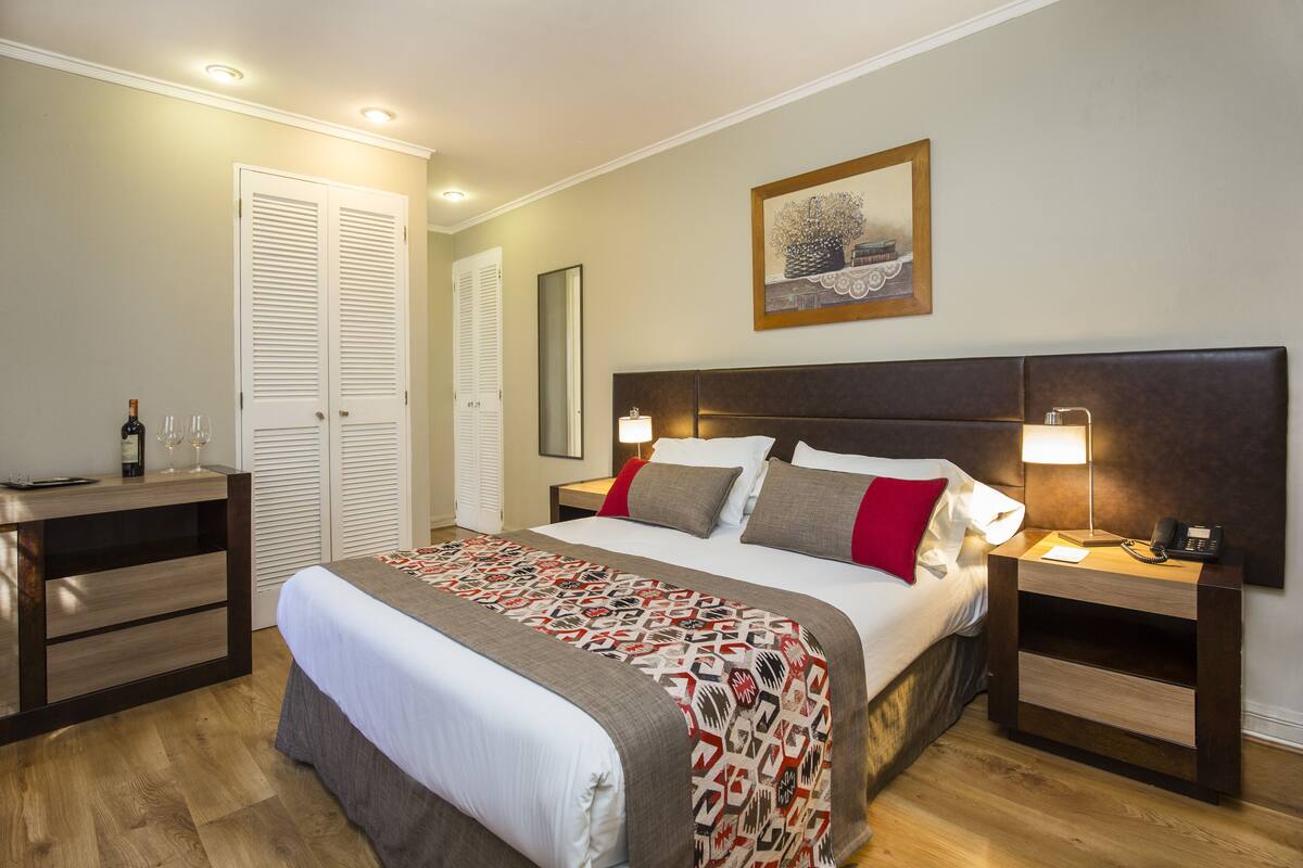 Une chambre d’hôtel bien rangée, avec un grand lit, des tables de chevet, une armoire et un tableau au mur.