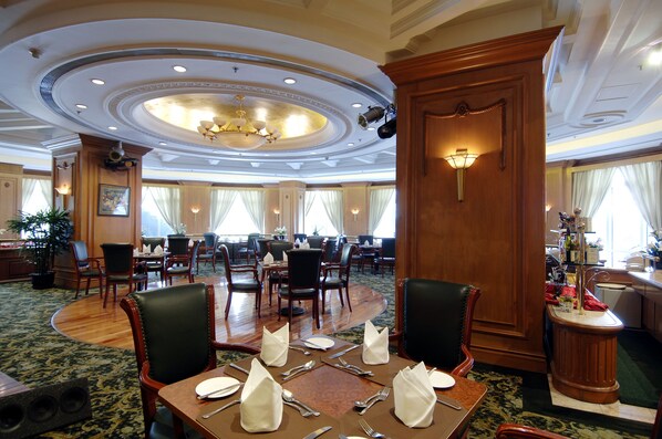 A hotel dining area with round tables set for four, wooden chairs, and a chandelier.