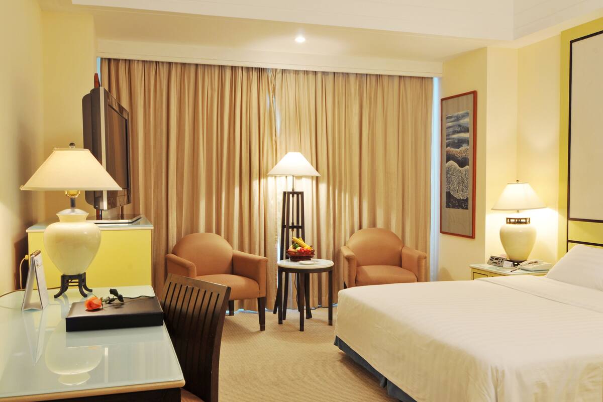 A hotel room with a bed, two armchairs, a small table with a fruit bowl, a desk with a lamp, and a painting on the wall.