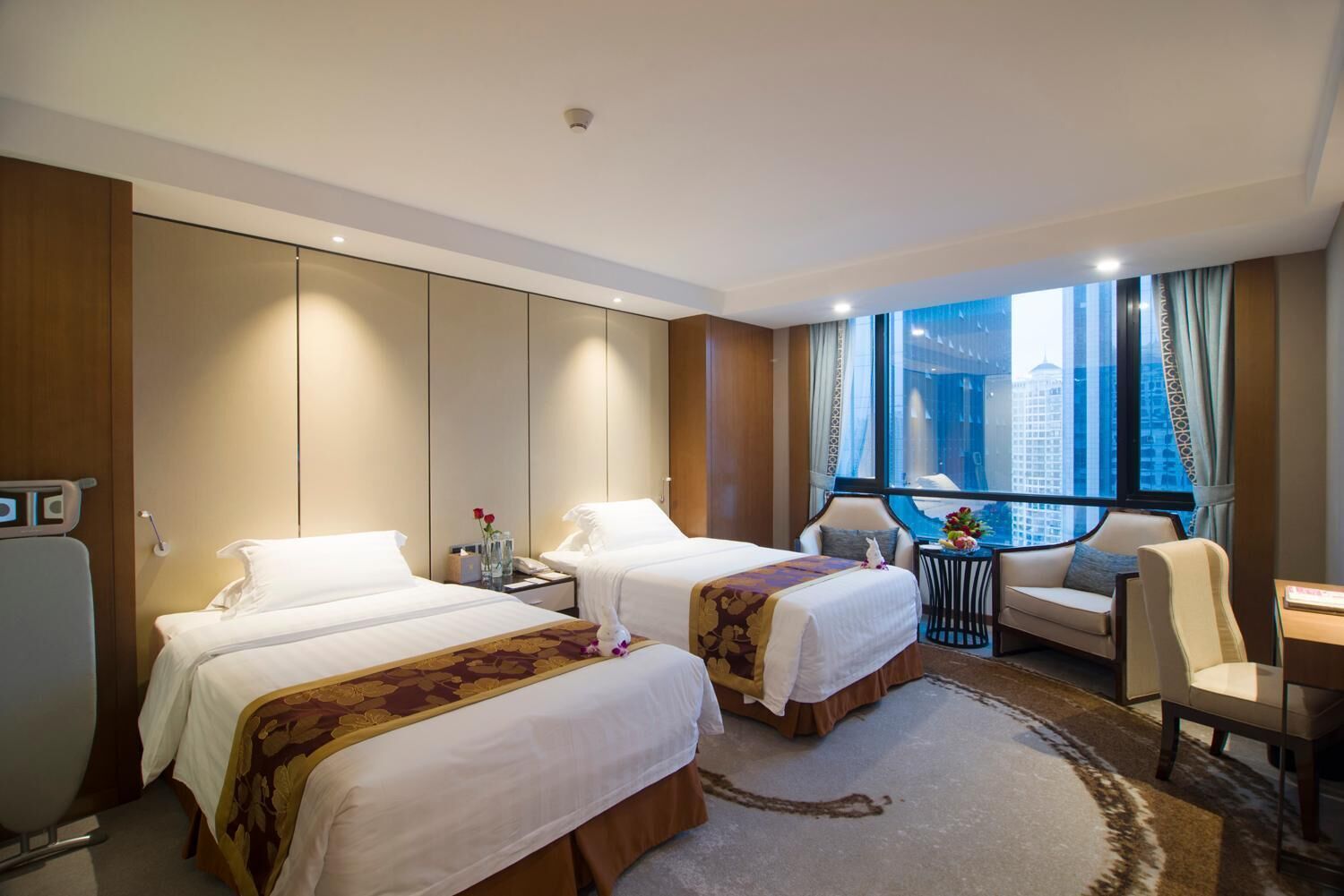 A hotel room with two beds, a desk, a chair, and a view of the city through large windows.