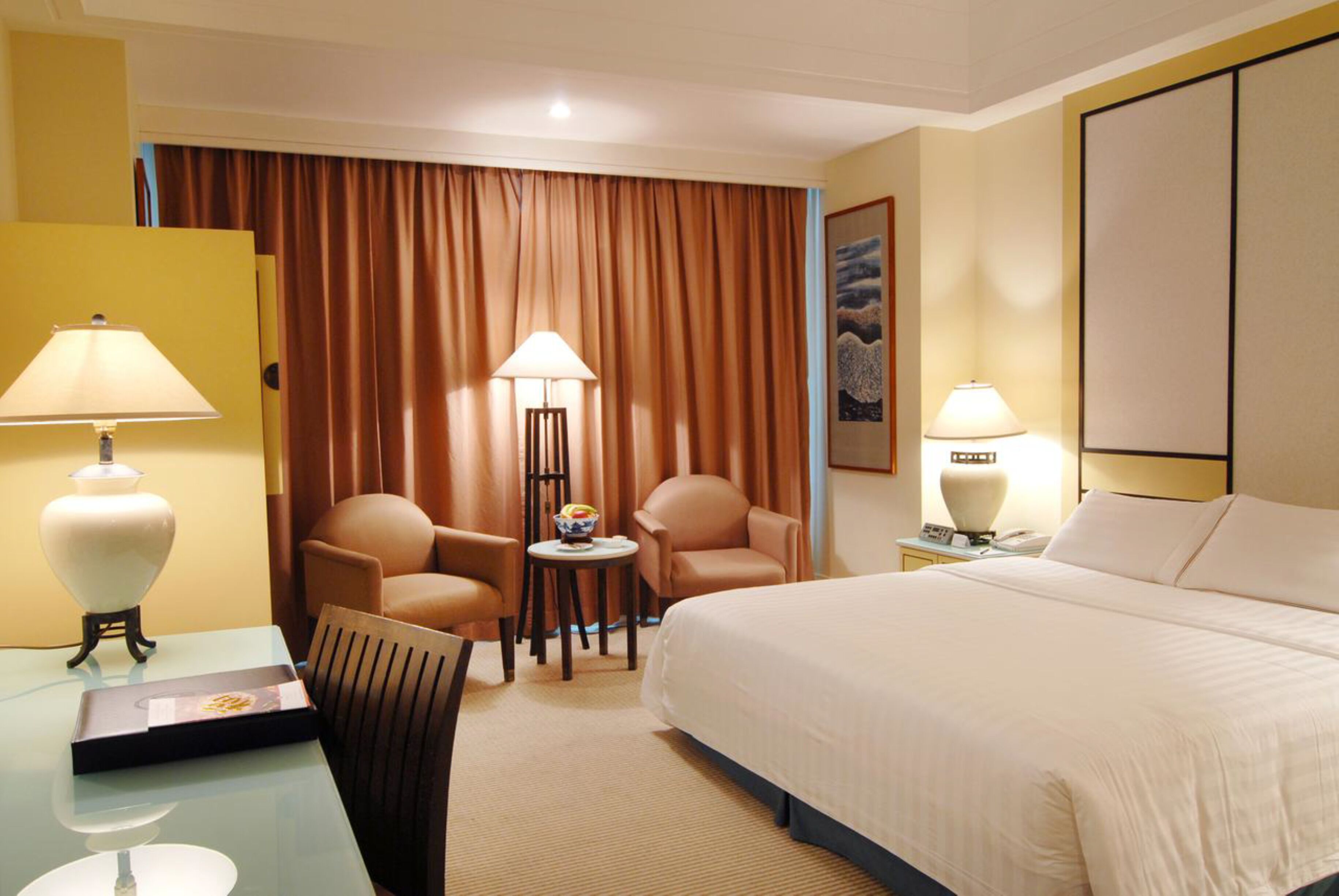 A hotel room with a bed, two armchairs, a small table with a bowl of fruit, two lamps, and a painting on the wall.