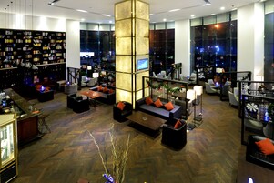 Un moderno vestíbulo de hotel con una zona de estar central, una amplia estantería y vistas a la ciudad a través de grandes ventanales.