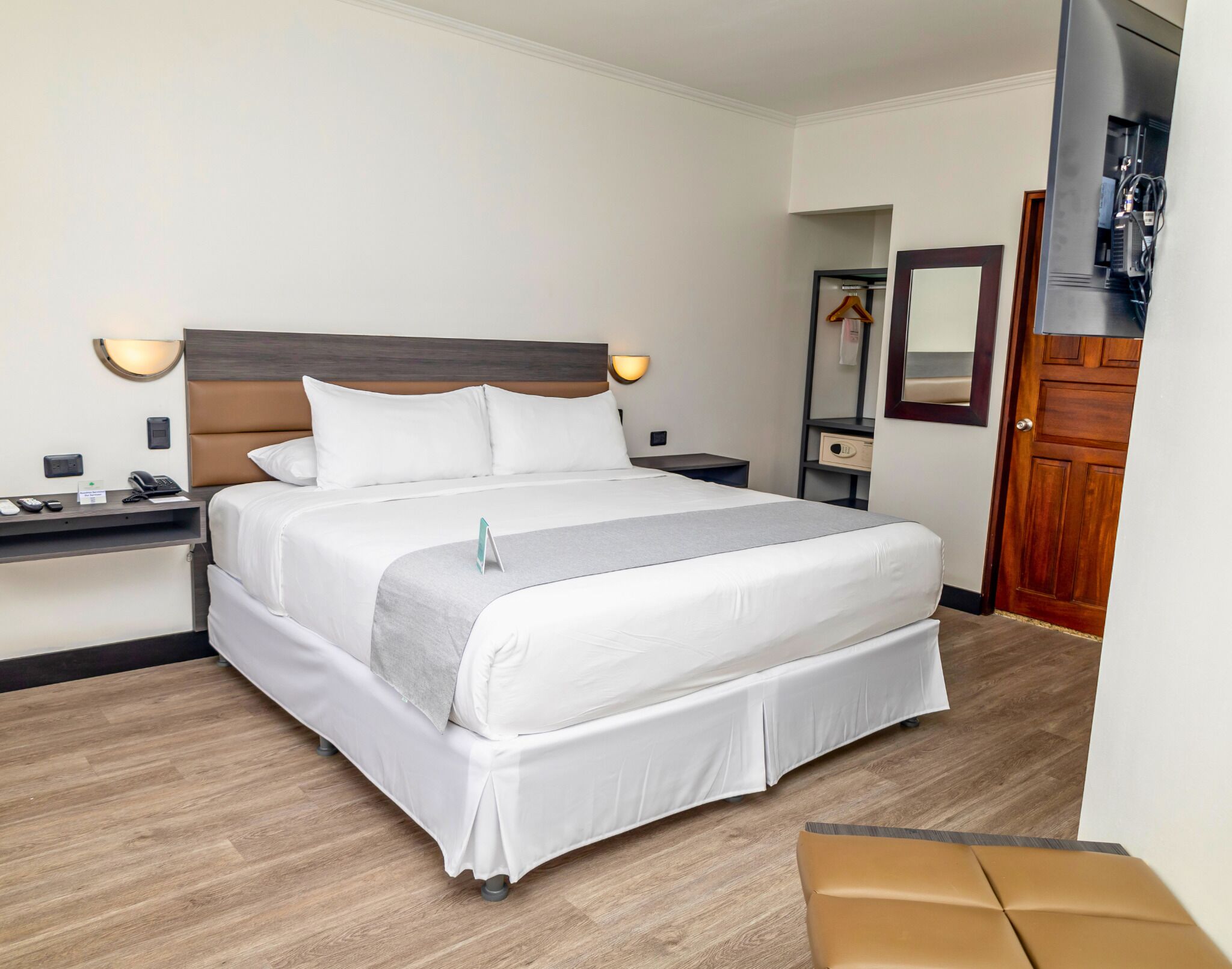 A hotel room with a bed, bedside tables, a nightstand, a wall-mounted TV, a mirror, and a wooden door.