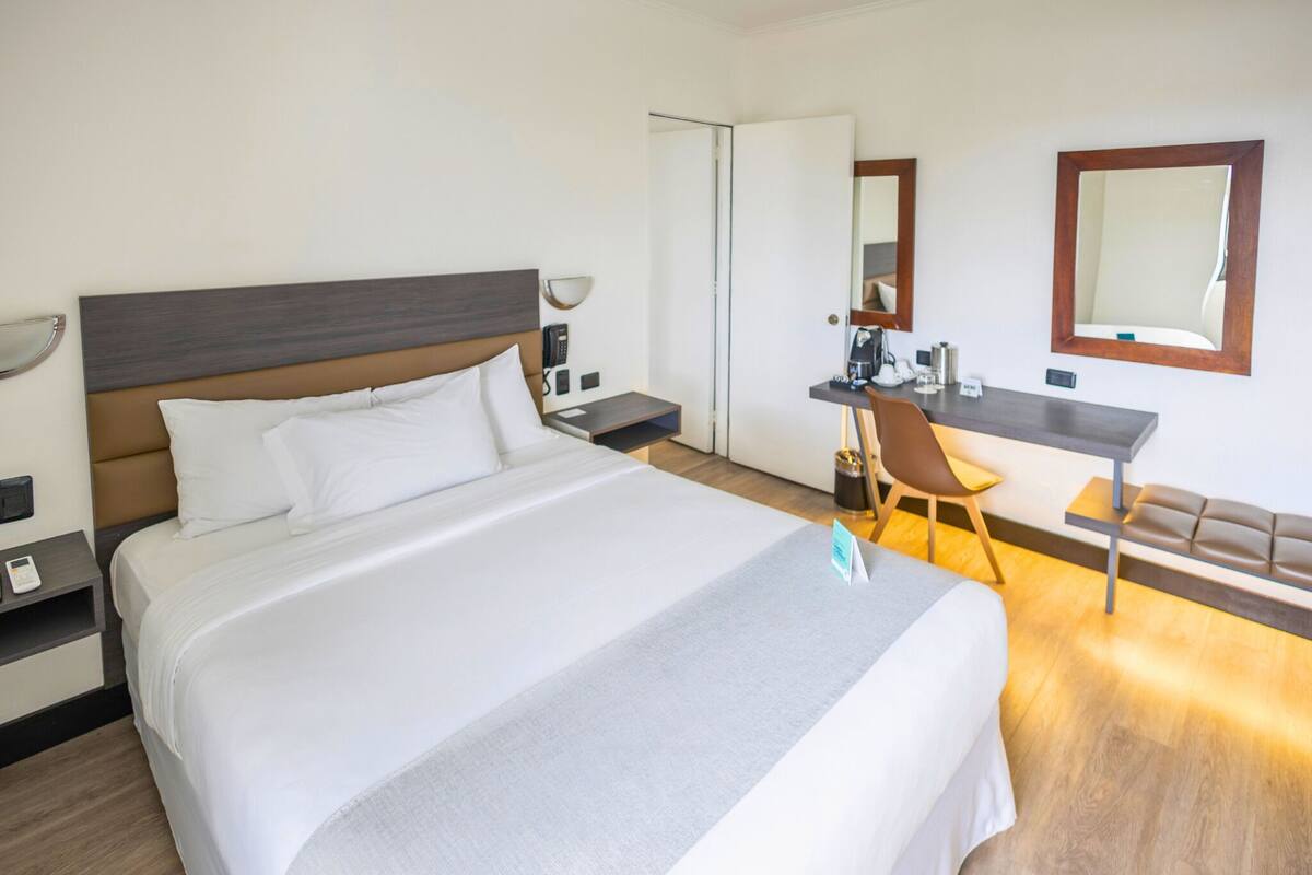 A hotel room with a large bed, a desk, a chair, a sofa, and a mirror.