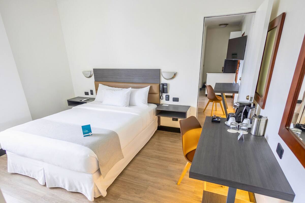 A hotel room with a bed, bedside table, chair, desk, and a mirror.