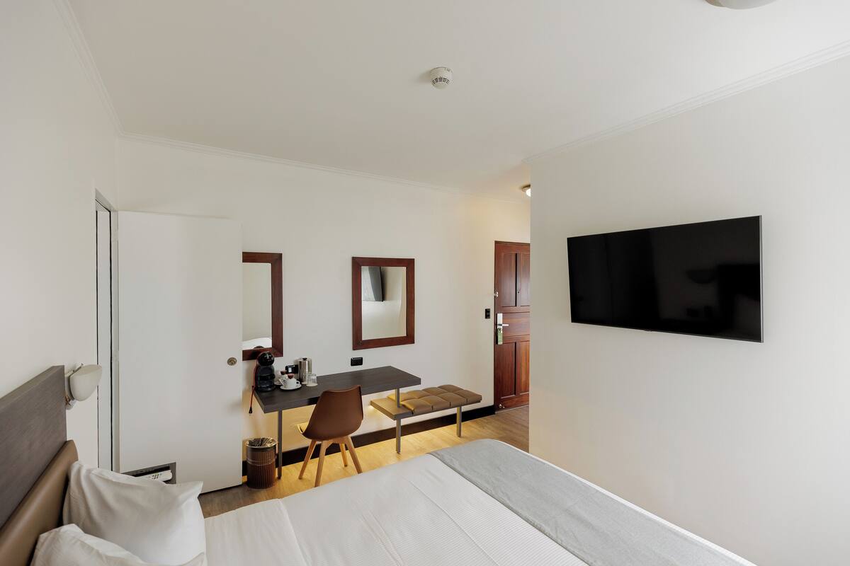 A hotel room with a bed, a desk with a chair, a television mounted on the wall, and a mirror above the desk.