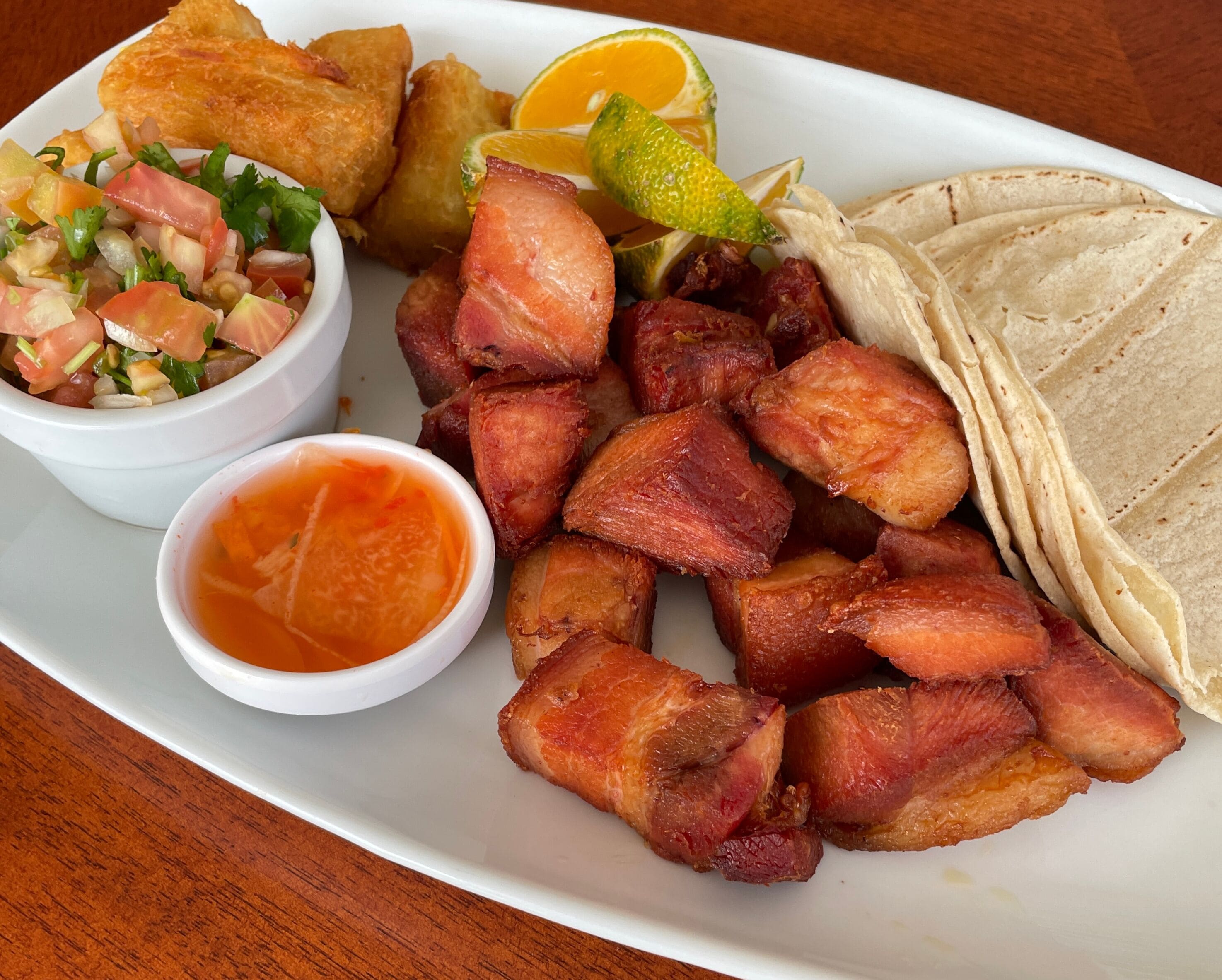 A plate with fried pork, a salad, a lime wedge, tortillas, and a small bowl of sauce.