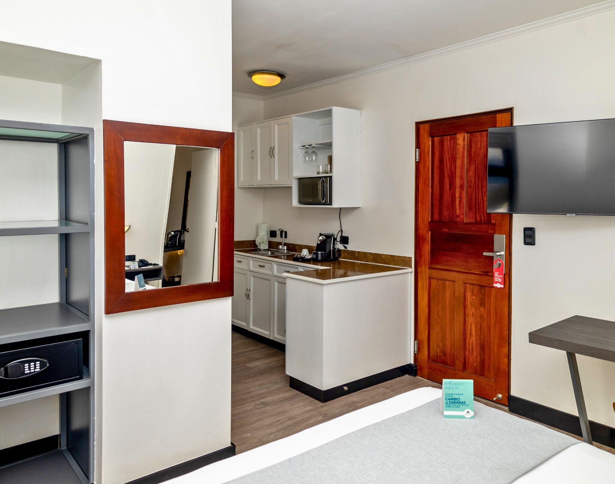 A compact hotel room with a kitchenette, a wooden door, a TV mounted on the wall, and a small table.