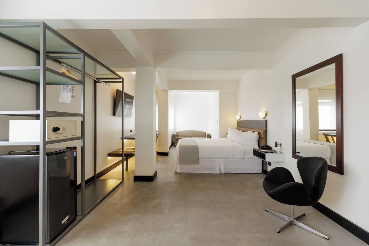 A hotel room with a bed, a desk, a chair, a mirror, and a minibar.