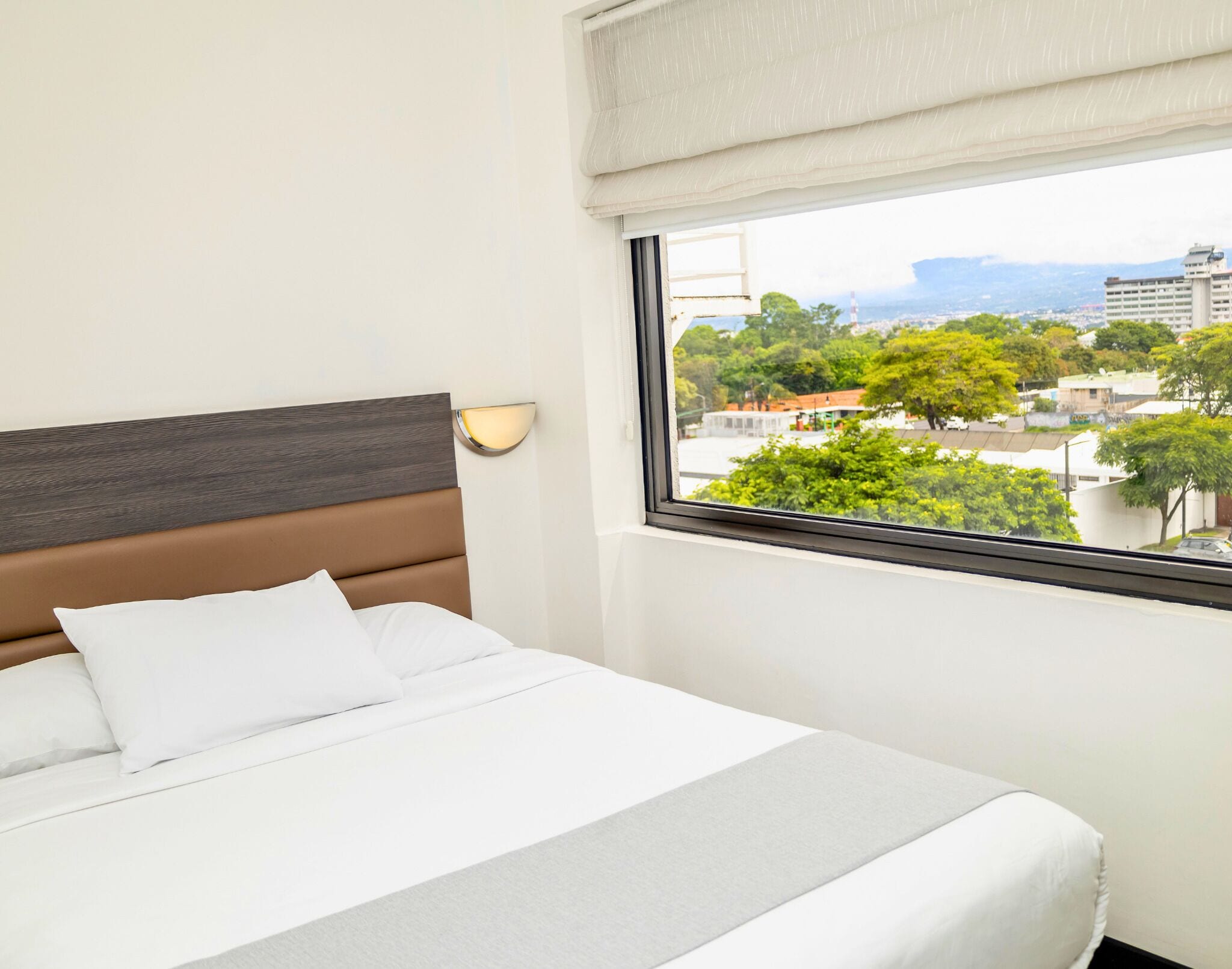Ein Hotelzimmer mit Bett, Kopfteil, Wandleuchte und Fenster mit Blick auf Stadt und Grünflächen.