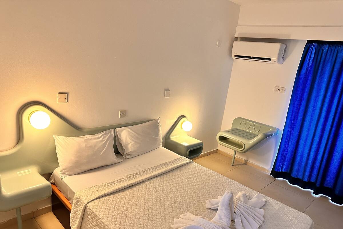 A neatly made bed with pillows, a bedside lamp, a small table, and a towel rack.