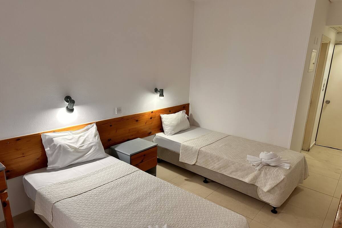 A hotel room with two single beds, wooden headboards, bedside tables, and a small nightstand.