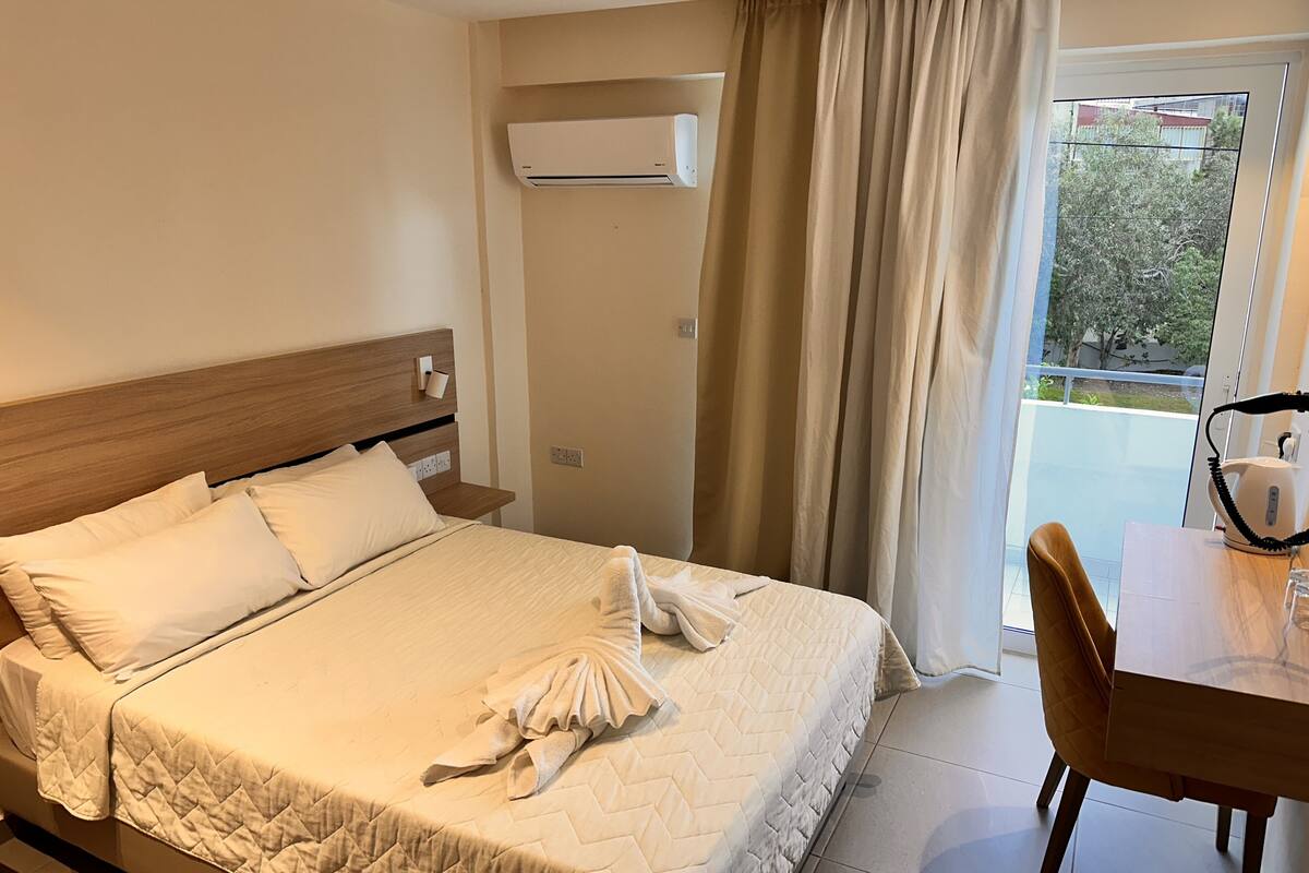 A hotel room with a bed, a wooden headboard, a bedside table, an air conditioning unit, a window with curtains, and a view of the outside.