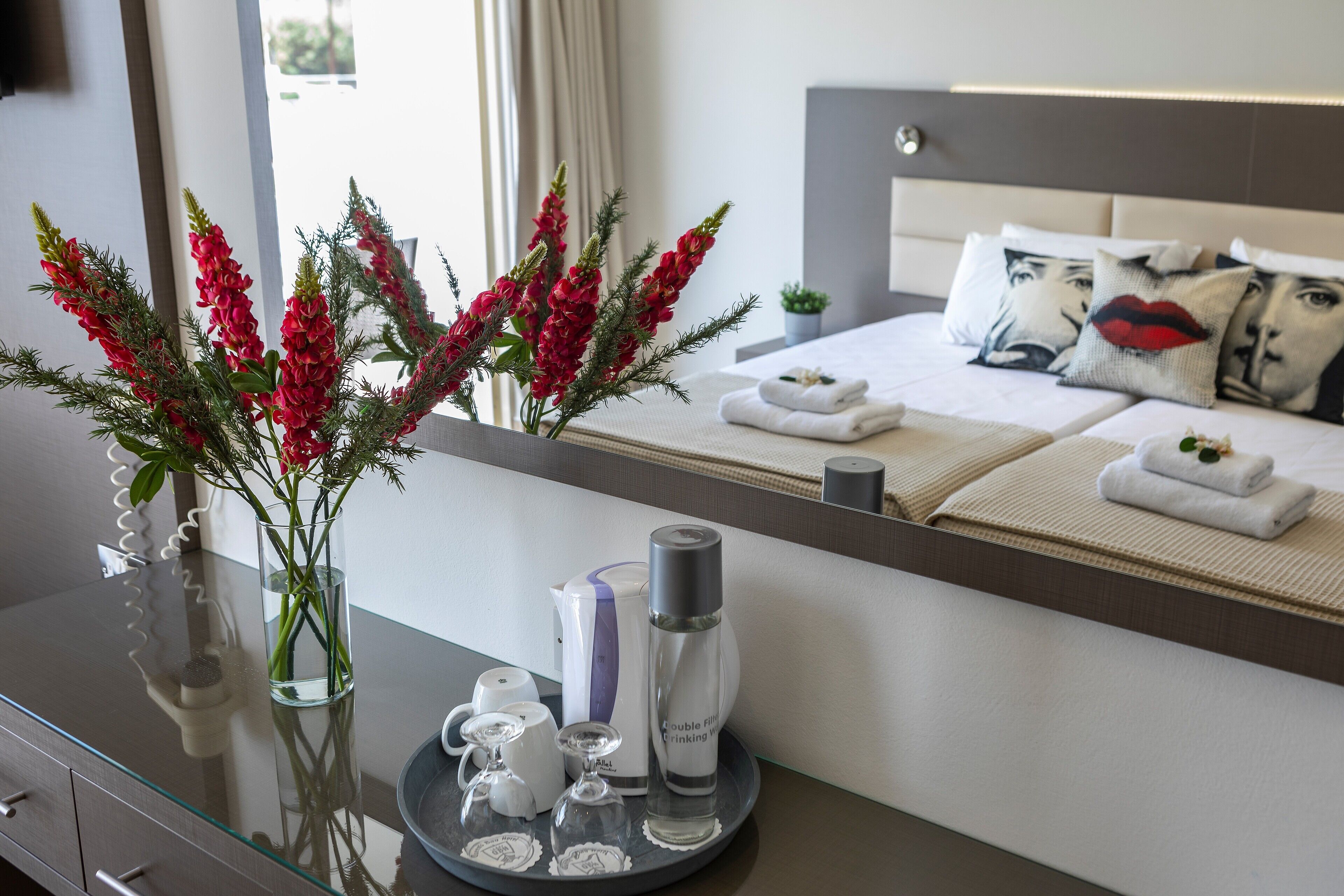 A hotel room with a bed, a bedside table with a vase of red flowers, a tray with toiletries, and a large mirror reflecting the room.