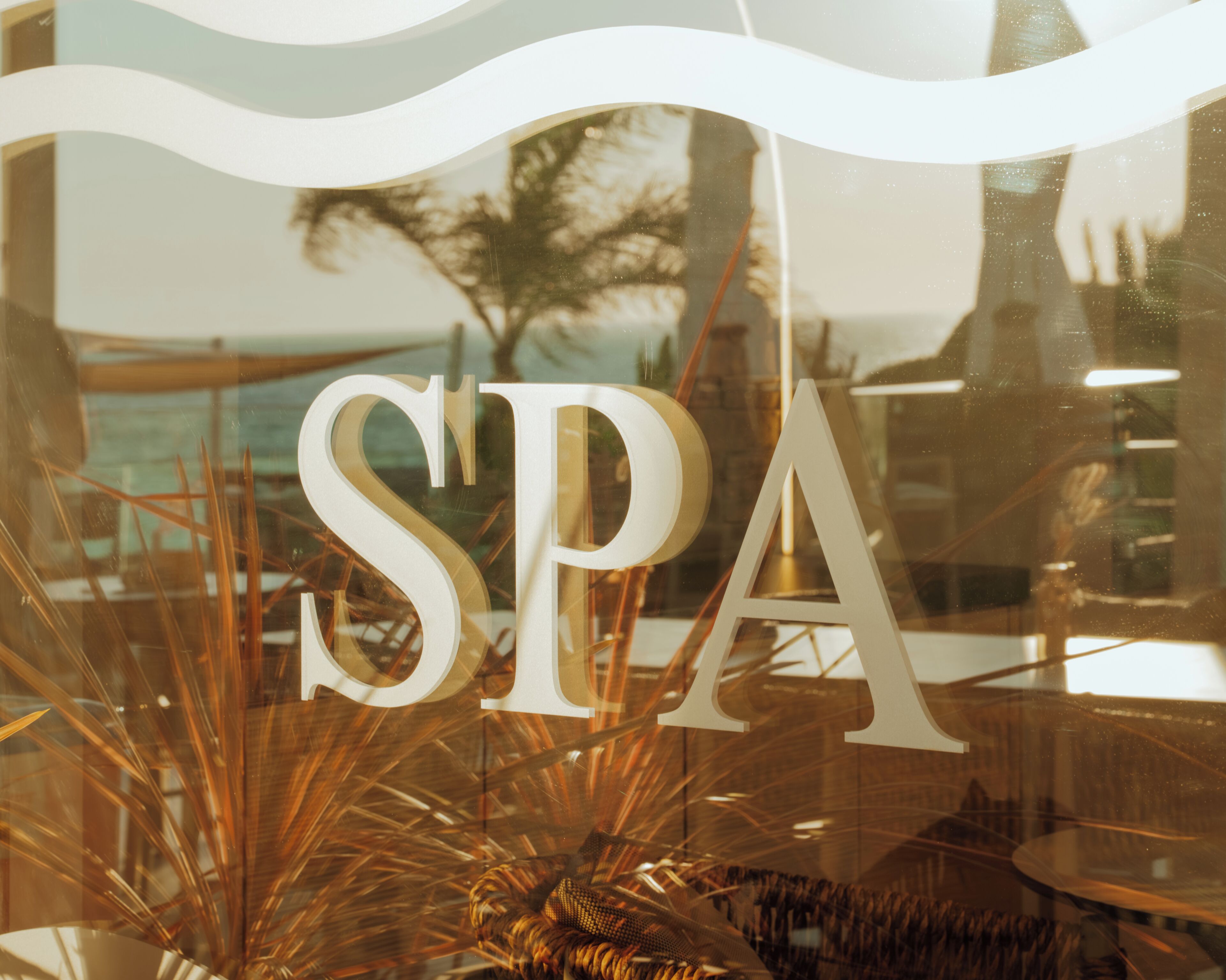 Een raam met de letters 'SPA' op de voorgrond, met uitzicht op een strand en palmbomen op de achtergrond.